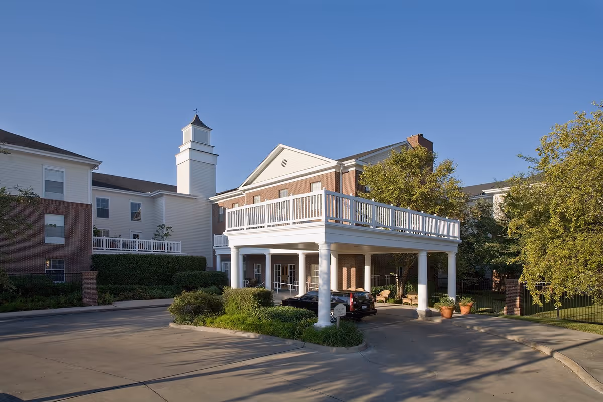 Exterior view of Belmont Village Senior Living West University building with a covered driveway entrance, white columns, brick and white siding, surrounded by greenery and trees under a clear blue sky.