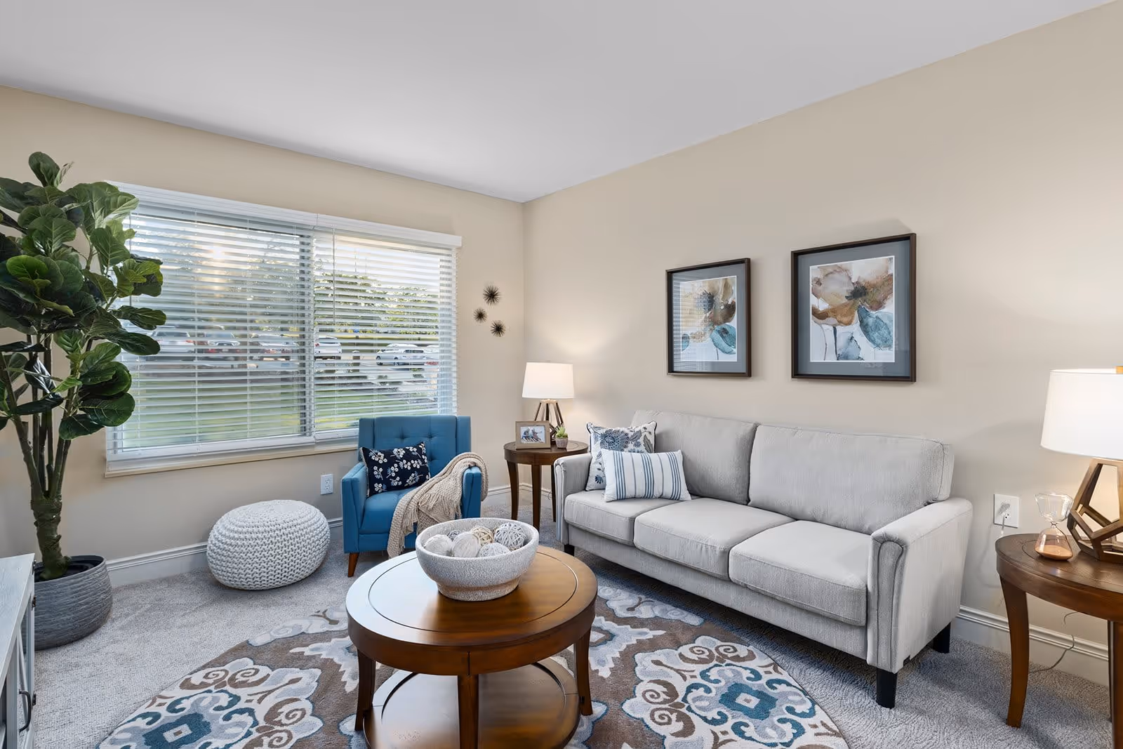 A cozy living room with a light gray sofa adorned with decorative pillows, a blue armchair with a throw blanket, a round wooden coffee table with a decorative bowl, a white knitted pouf, a large green potted plant near a window with blinds, two framed floral artworks on the beige wall, and two wooden side tables each with a lamp.
