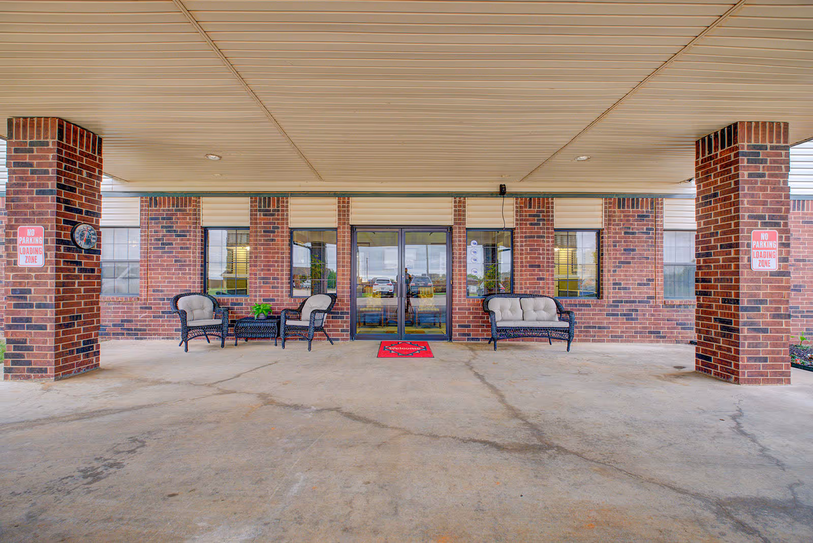 Covered entrance area of a brick building with two cushioned chairs and a small table on the left, and a cushioned bench on the right. The entrance has glass double doors and windows on either side. Two brick pillars have 'No Parking Loading Zone' signs attached.