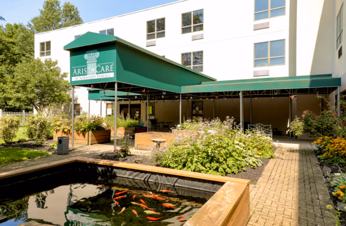Outdoor garden area at AristaCare at Norwood Terrace featuring a covered patio with green awnings, raised wooden garden beds with plants and flowers, a small pond with orange fish, and a multi-story building in the background.