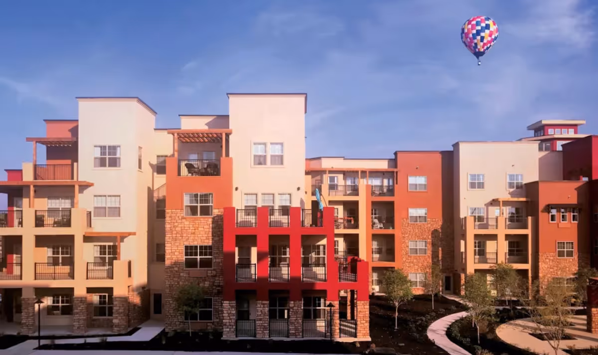 Front facade of a multi-story senior living apartment building with balconies and a hot air balloon in the sky.