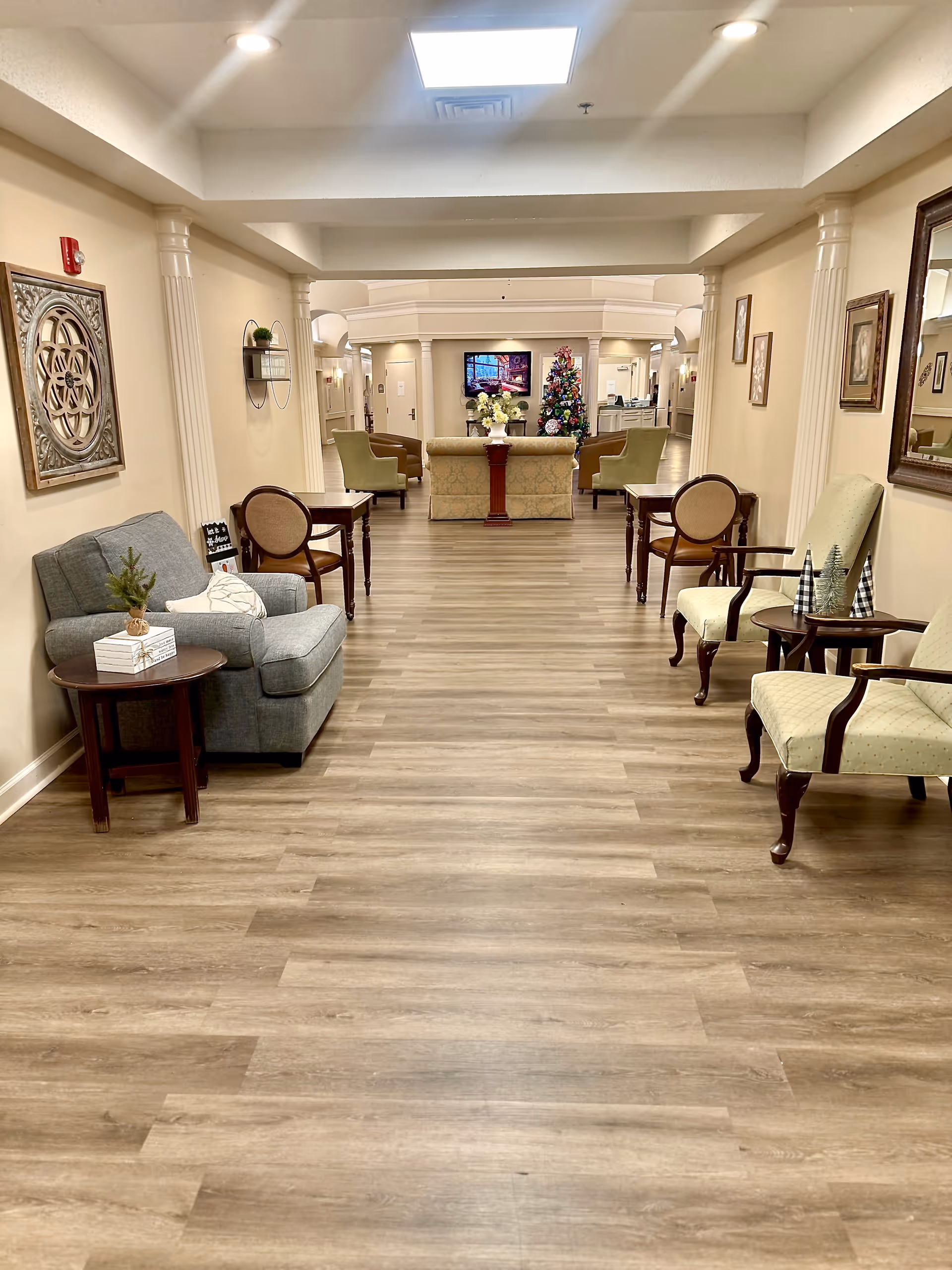 A spacious and well-lit senior living facility common area with wooden flooring, beige walls, and white columns. The room is furnished with various chairs and small tables on both sides of the hallway. In the background, there is a decorated Christmas tree and a television mounted on the wall above a floral arrangement on a wooden pedestal.