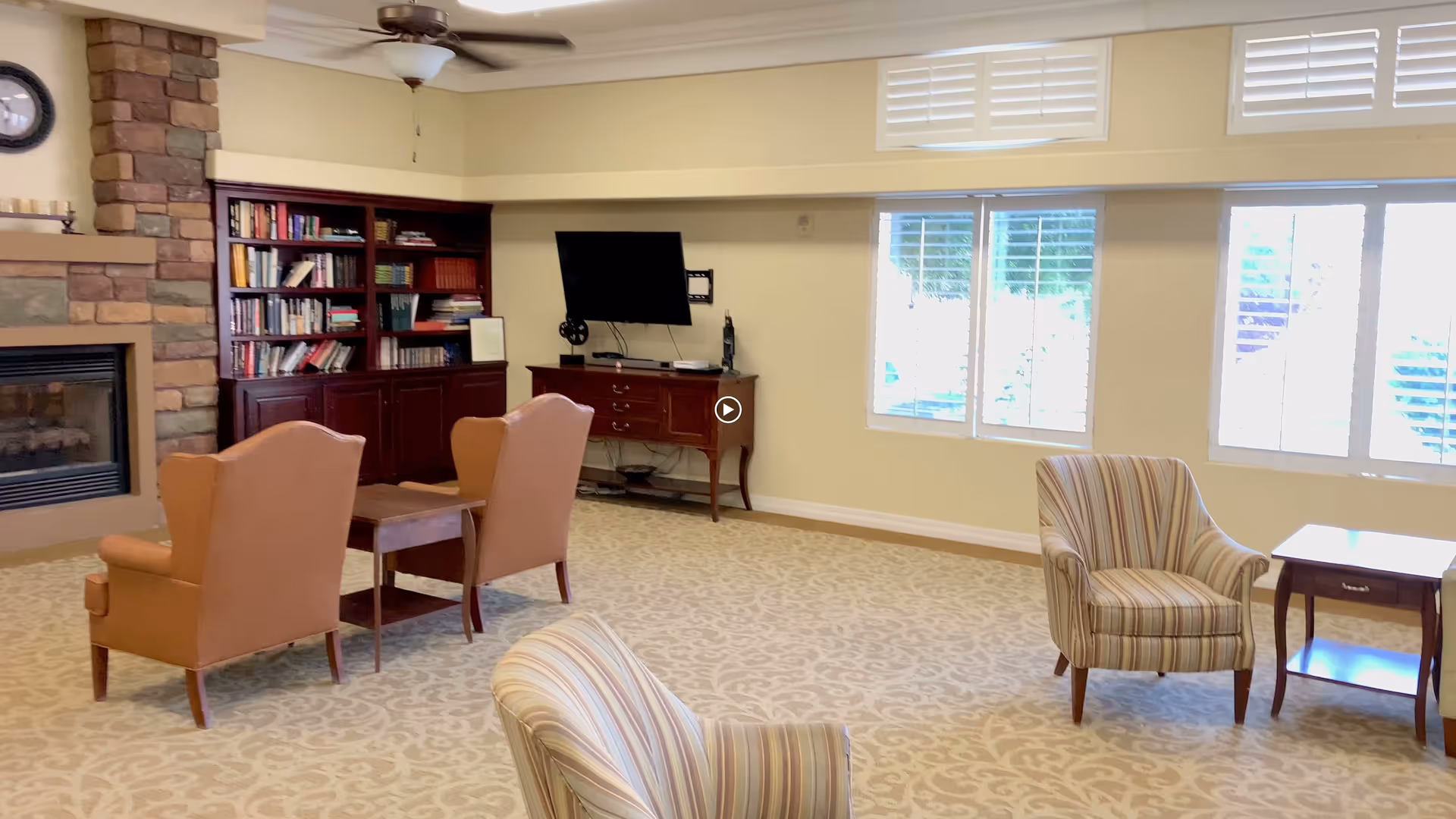 A bright and spacious living room with beige patterned carpet, two brown armchairs facing a wooden coffee table, a striped armchair near a small wooden side table, a stone fireplace on the left, a wooden bookshelf filled with books, a flat-screen TV mounted on the wall above a wooden console, and large windows with white shutters letting in natural light.