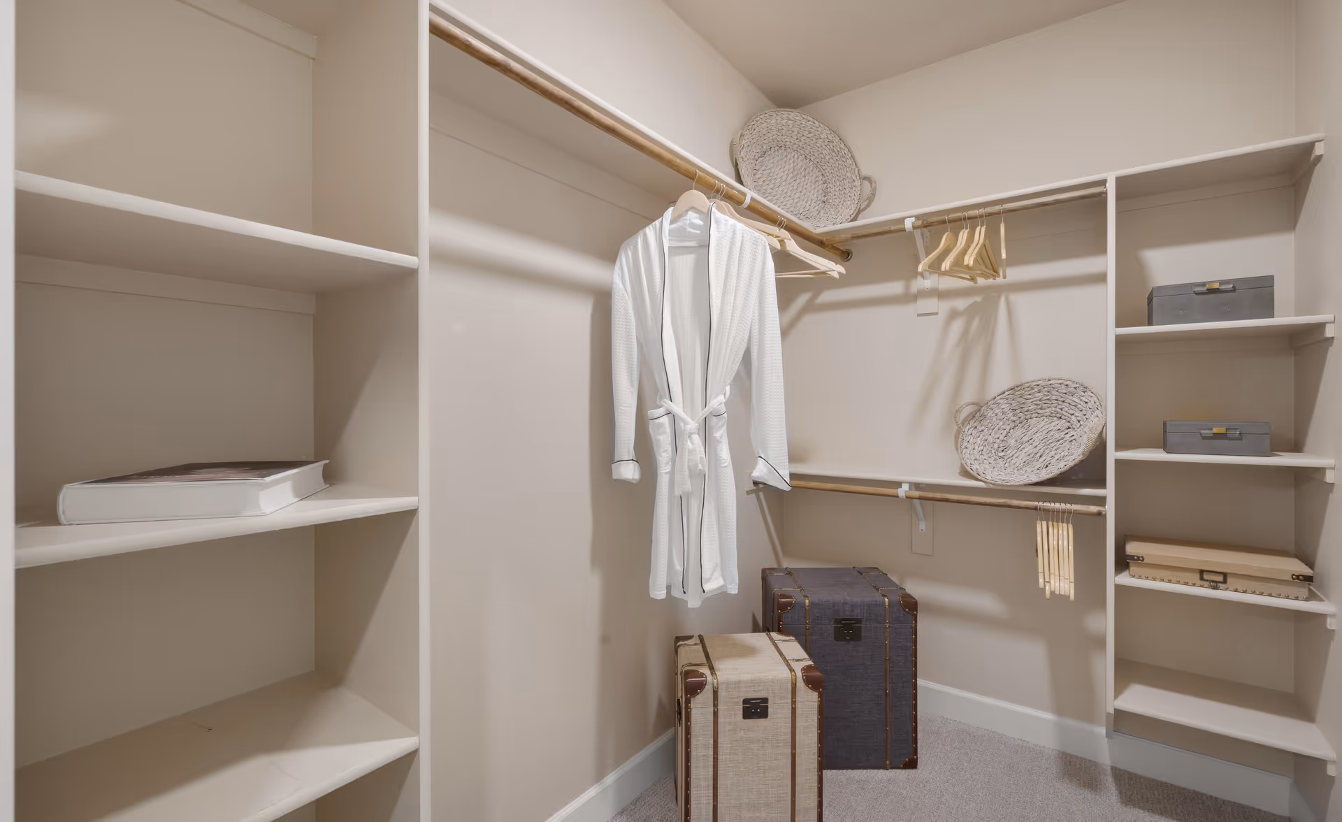 A walk-in closet with built-in shelves and hanging rods. The closet contains a white robe hanging on a rod, two decorative baskets on shelves, and two vintage-style suitcases on the floor. The walls are painted beige and the carpet is light-colored.