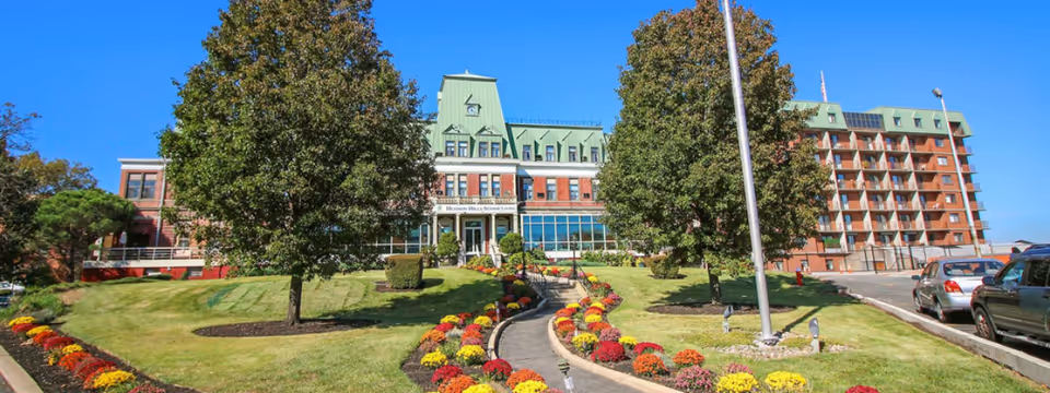 Front exterior view of a senior living facility with a large brick building featuring green roofs, a central entrance with steps, and a landscaped garden with colorful flowers and trees along a curved pathway. Several cars are parked on the right side near the building.