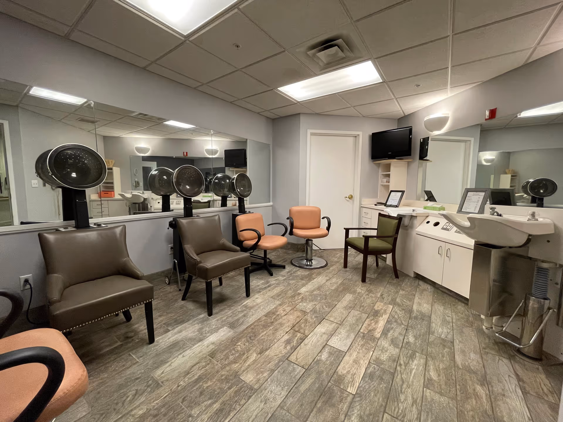 Salon-style room with multiple hooded hair dryers, various chairs, large mirrors, a sink and cabinets.