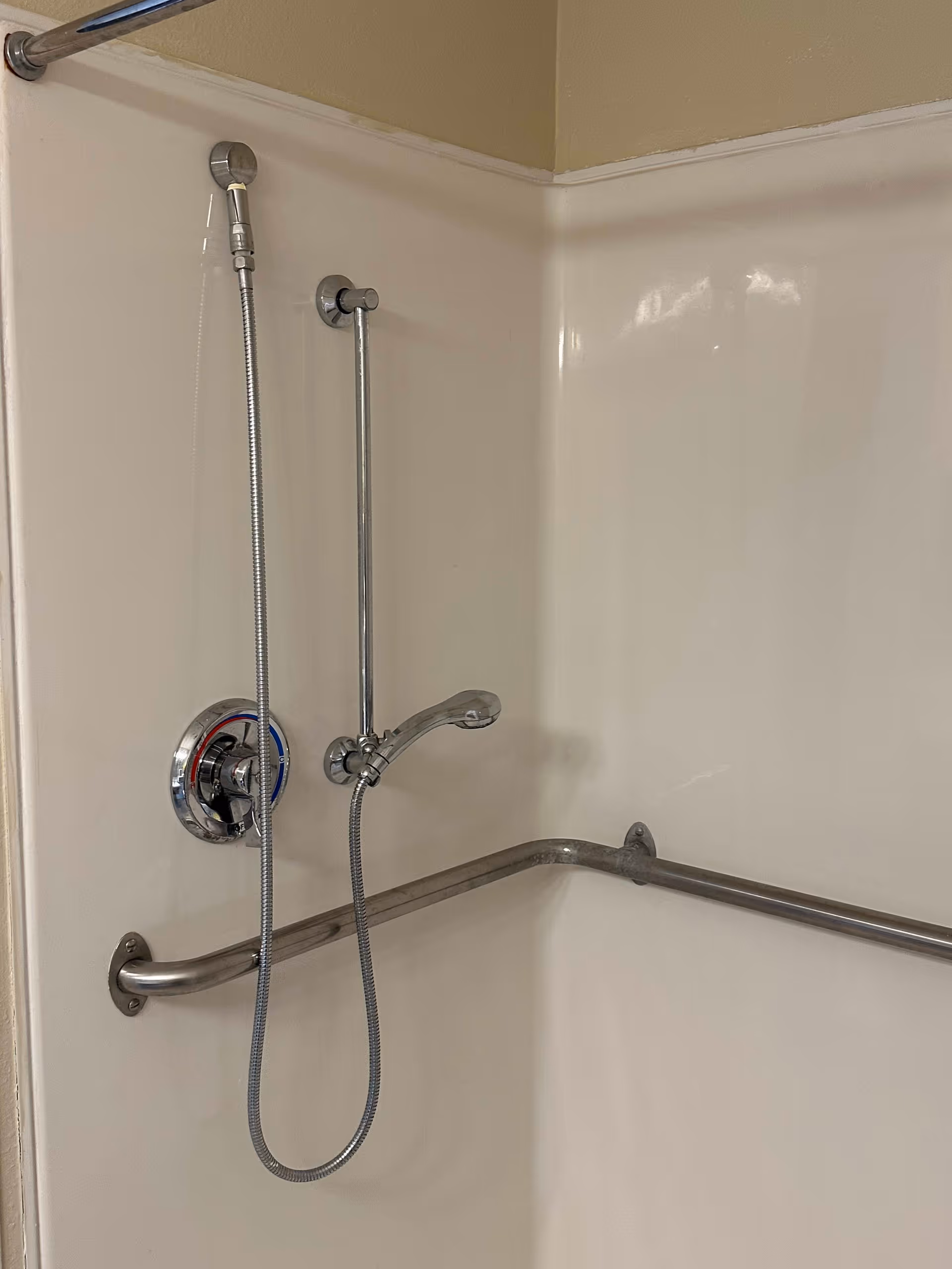 A shower area with a handheld showerhead attached to a vertical sliding bar, a faucet, and a horizontal grab bar mounted on the wall for support. The walls are smooth and light-colored.