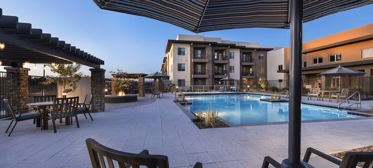 Outdoor patio area at Inspira at Arrowhead featuring a swimming pool surrounded by lounge chairs and tables with umbrellas. The area includes a pergola with string lights and a fire pit, with a multi-story residential building in the background under a clear evening sky.