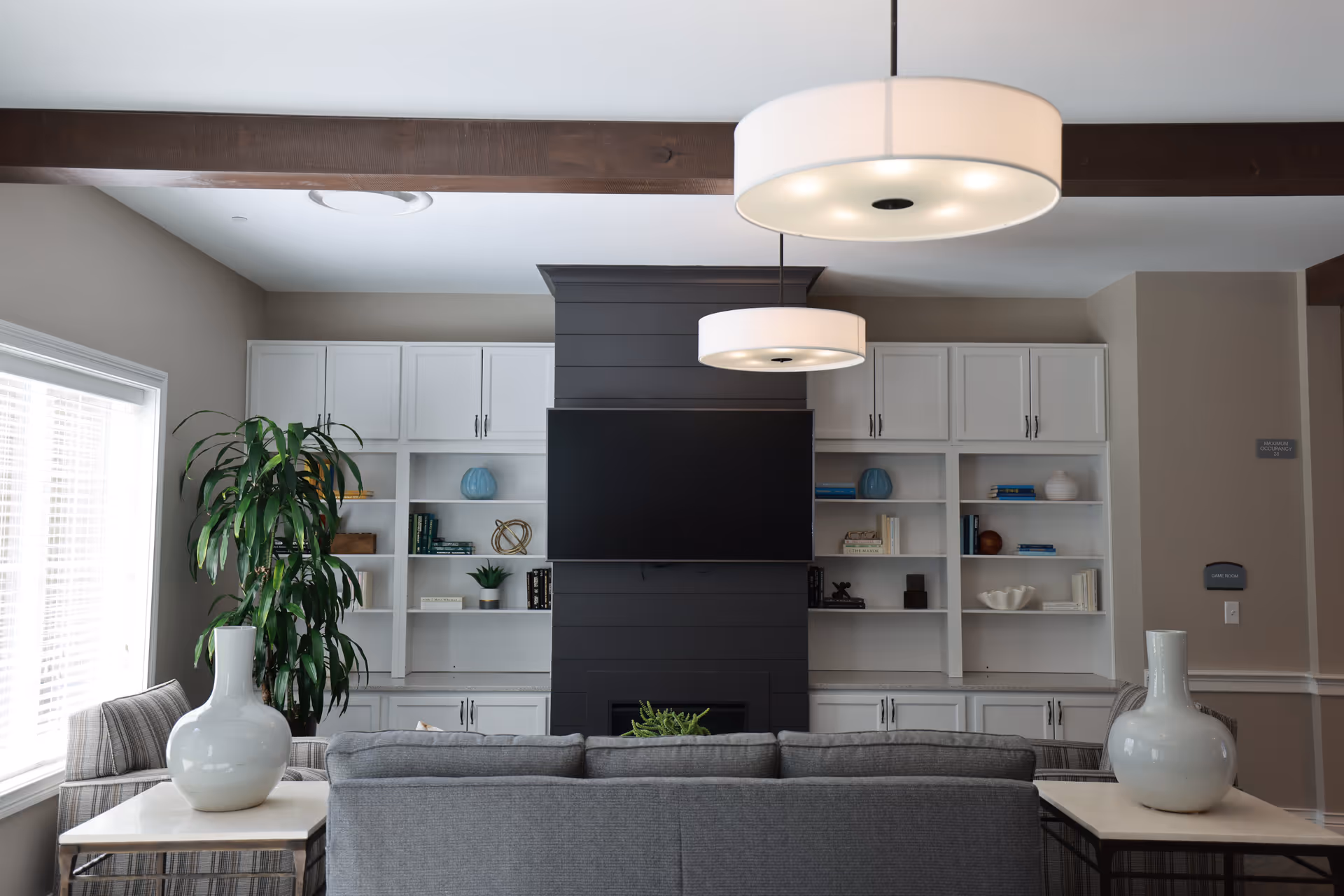 A modern living room with a gray sofa facing a black shiplap wall-mounted flat screen TV. The wall behind the TV has white built-in shelves and cabinets decorated with books, vases, and small plants. Two large white pendant lights hang from the ceiling, and there is a large window with blinds on the left side. Two white vases are placed on side tables next to the sofa, and a tall green plant is positioned near the window.