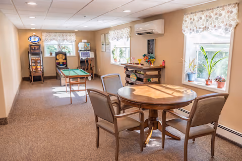 Bright communal activity room with a round wooden table and chairs, a small pool table, arcade games, and windows with potted plants.