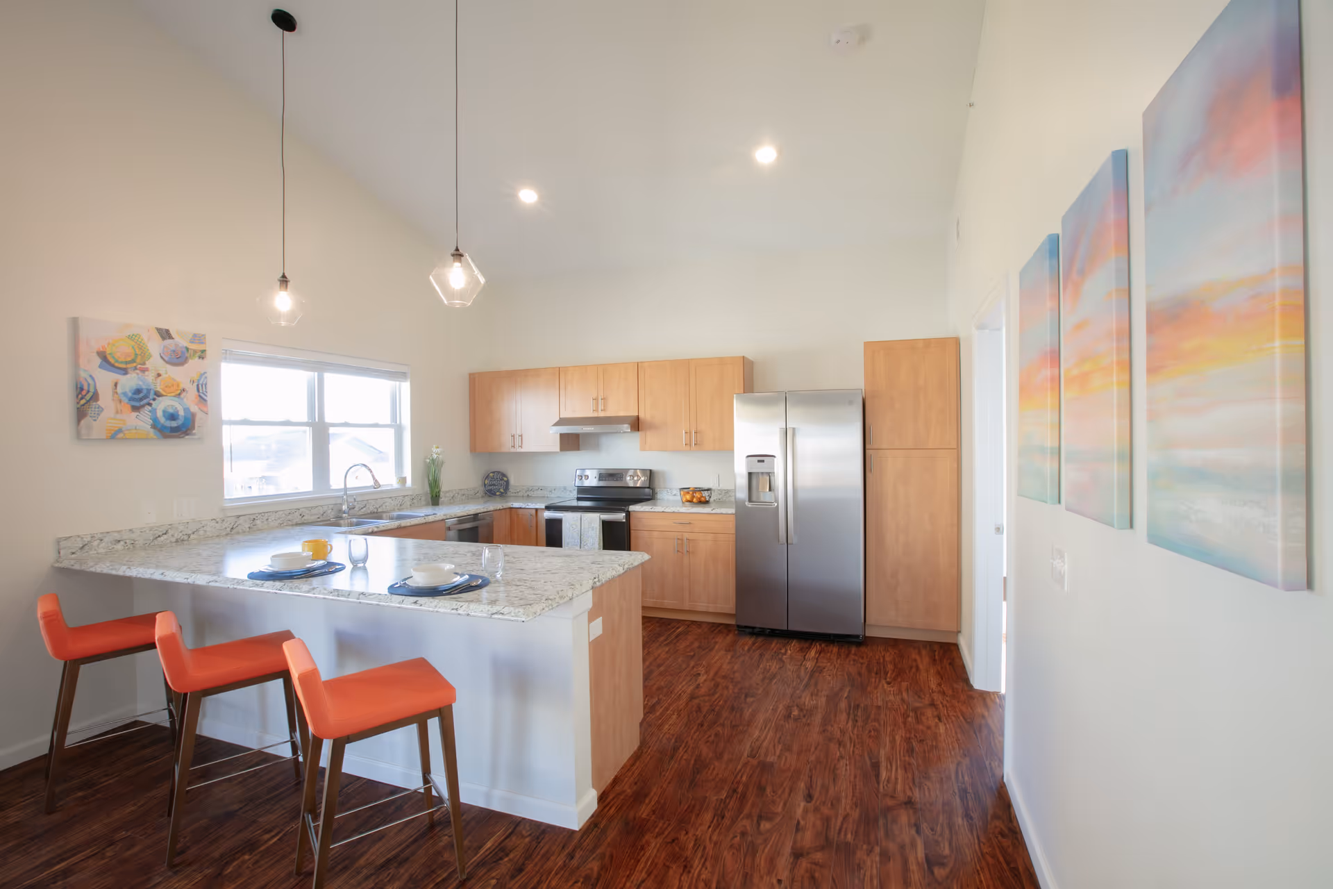 Bright modern kitchen with wooden cabinets, stainless steel refrigerator and stove, granite countertops, two orange bar stools at the kitchen island, pendant lights hanging from the ceiling, and colorful artwork on the walls.