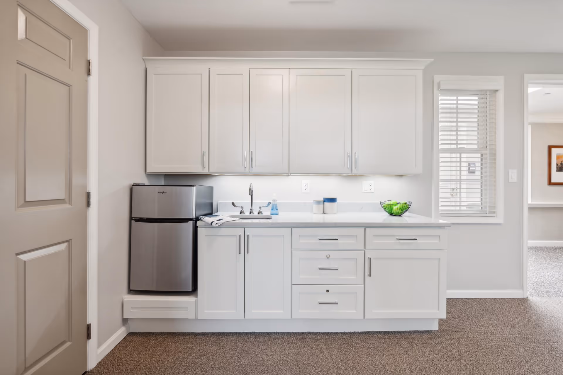 A small kitchenette area with white cabinets, a stainless steel mini refrigerator, a sink with a faucet, and a countertop with a green bowl and two small containers. There is a window with white blinds to the right and a beige door to the left.