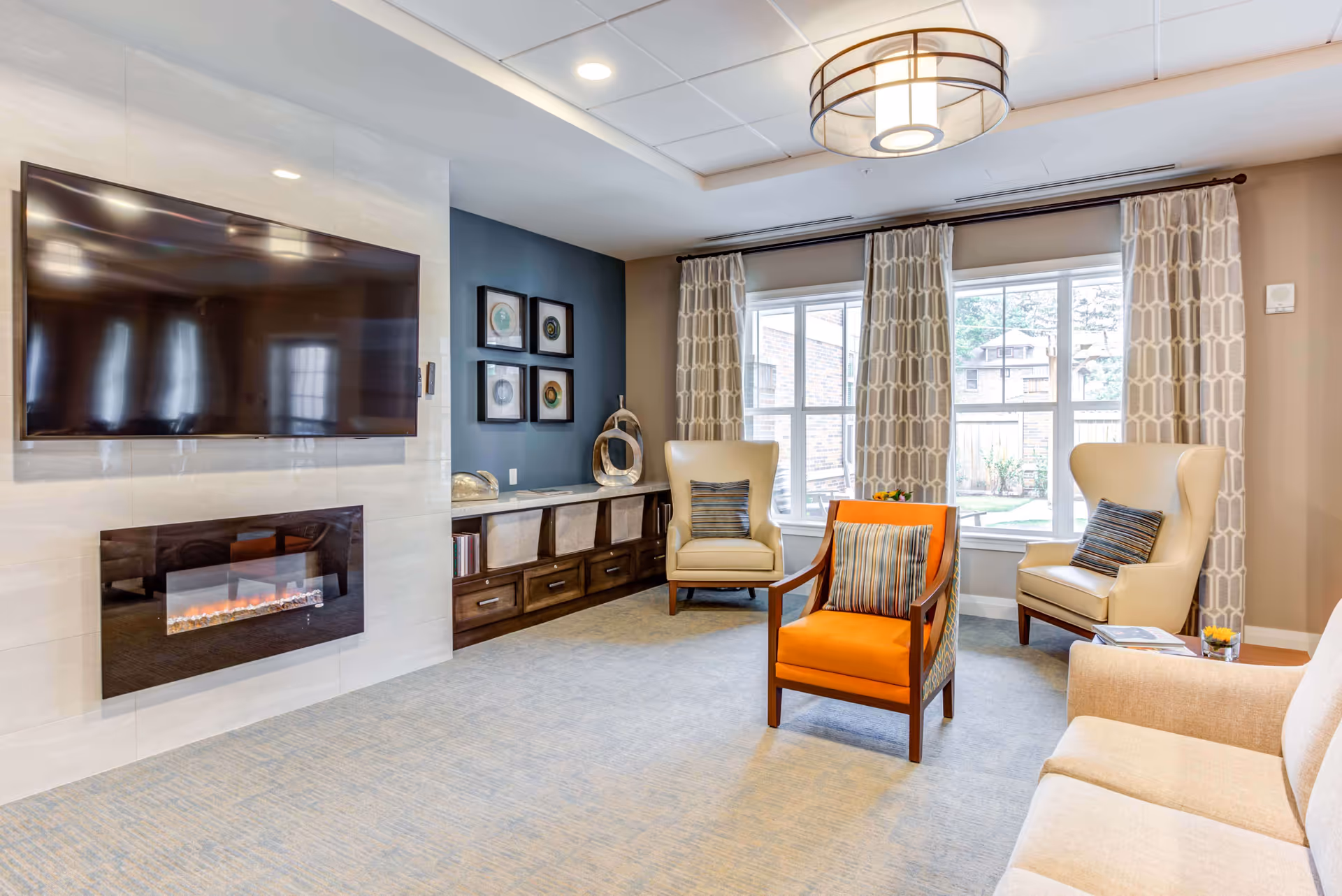 A bright and cozy living room with a large flat-screen TV mounted above a modern electric fireplace. The room features a beige sofa, two cream-colored armchairs with striped pillows, and an orange armchair with a striped pillow. There is a large window with patterned curtains letting in natural light, and a wooden cabinet with decorative items and framed artwork on the wall.
