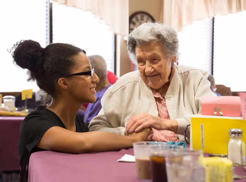 A young caregiver smilingly holds hands with an elderly woman seated at a table in a communal dining room.