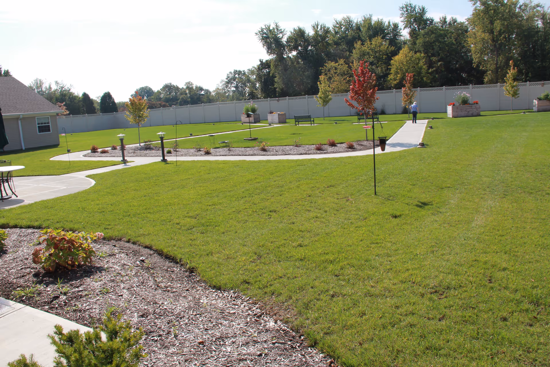 A spacious outdoor garden area with green grass, small trees, and a paved walkway. There are benches and raised flower beds along the path, and a person walking in the distance near a white fence. The sky is clear and sunny.