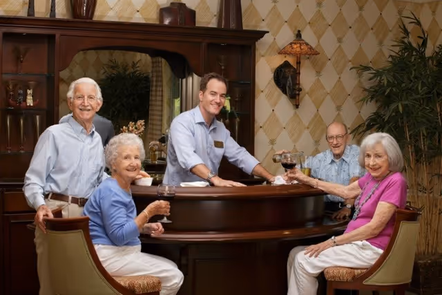Four elderly people and a staff member gathered around a wooden bar counter in a cozy room with patterned wallpaper and decorative plants. The elderly individuals are seated on bar stools, holding glasses of red wine, smiling and engaging with the staff member who is standing behind the bar.