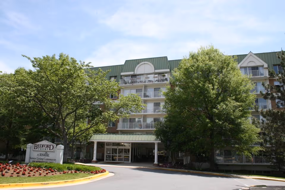 Exterior view of Bedford Court, a senior living community building with multiple floors, balconies, and a green roof. The entrance is covered and surrounded by trees and landscaping with flowers. A sign near the entrance reads 'Bedford Court A Sunrise Senior Living Community.'