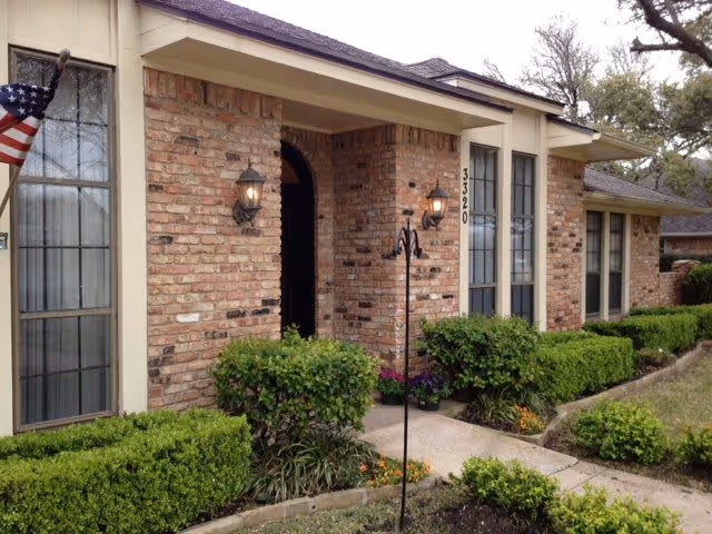 Exterior view of a single-story brick residential building with large windows, two wall-mounted lantern lights flanking the entrance, neatly trimmed bushes and plants along the walkway, and an American flag on the left side.