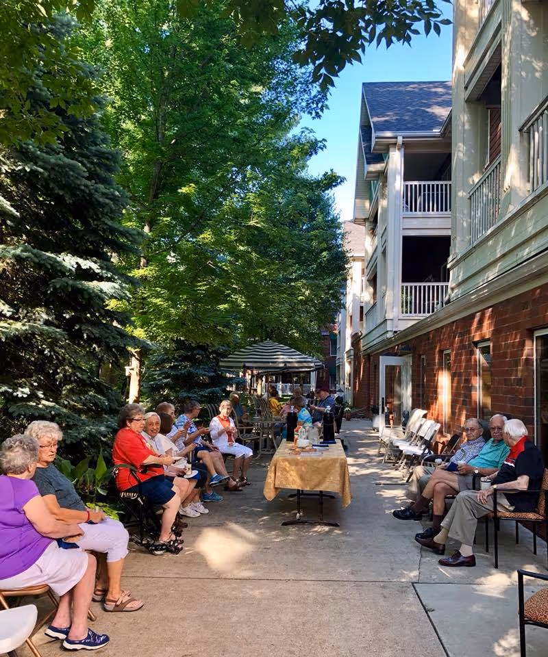 A group of elderly people sitting outdoors along a paved walkway next to a building with balconies. They are seated on chairs on both sides of the walkway, some engaging in conversation. There is a table with a yellow tablecloth in the middle of the walkway with items on it. The area is shaded by large green trees and there is a striped umbrella further down the path.
