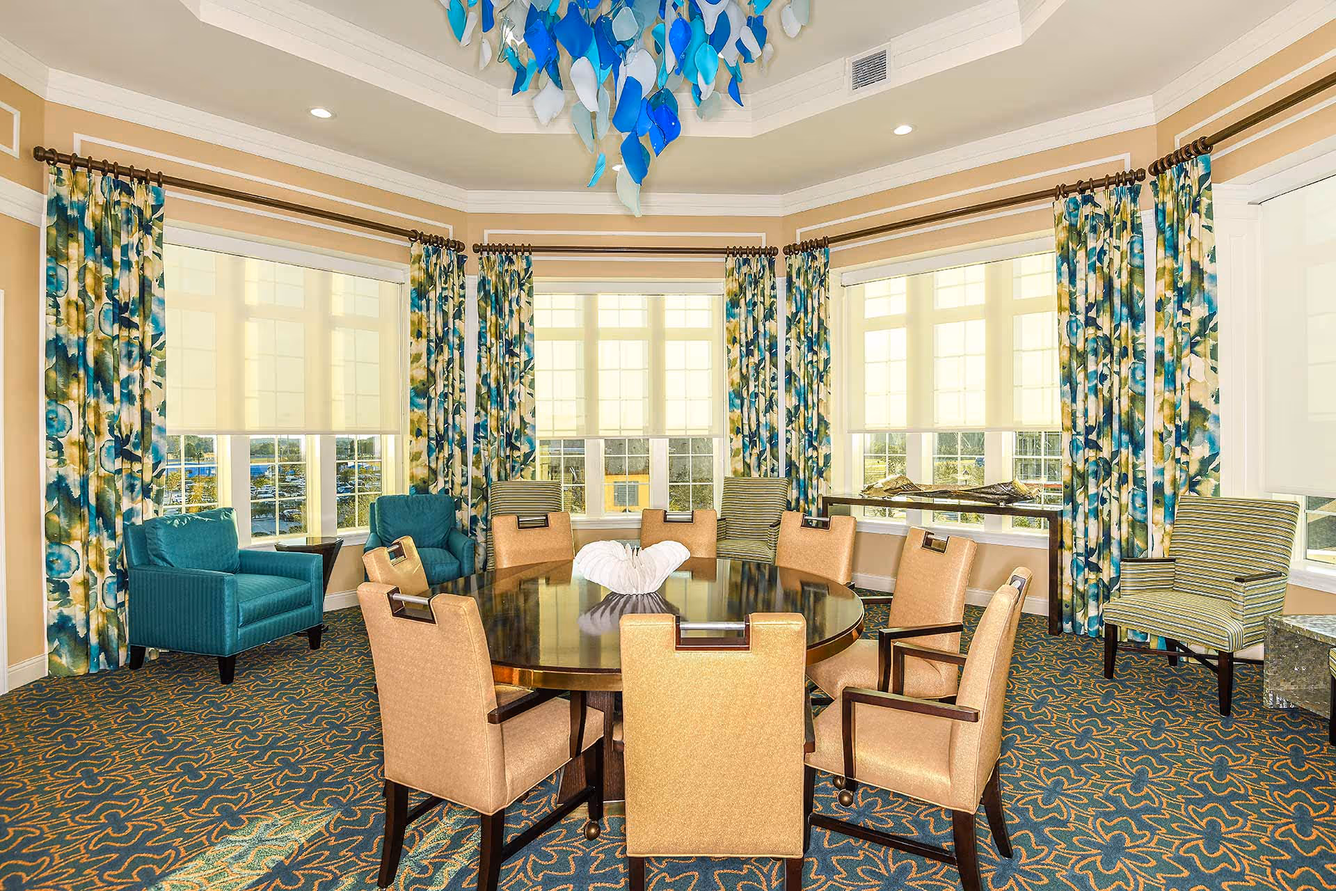 A bright and elegant room with a large round wooden table surrounded by eight beige upholstered chairs. The room features large windows with floral patterned curtains in shades of blue, green, and yellow. There are two teal armchairs and two striped armchairs placed near the windows. The ceiling has a decorative light fixture with blue and white glass elements. The carpet has a blue and orange floral pattern.