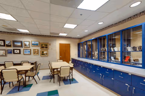 Community dining/activity room with several tables and chairs, framed pictures on one wall and blue display cabinets along the opposite wall.