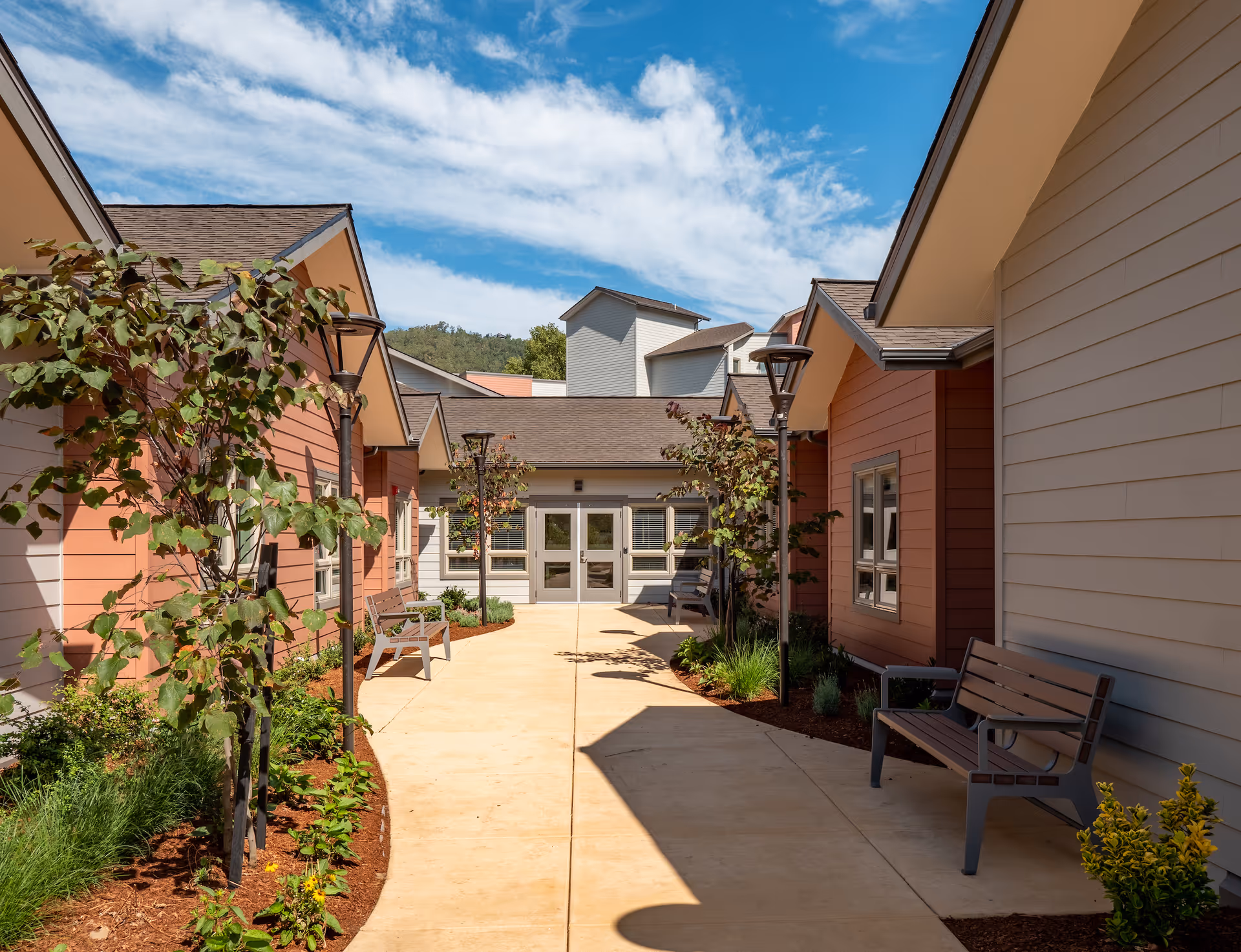 Outdoor walkway between buildings at a senior living community with benches, small trees, and plants along the path under a partly cloudy blue sky.