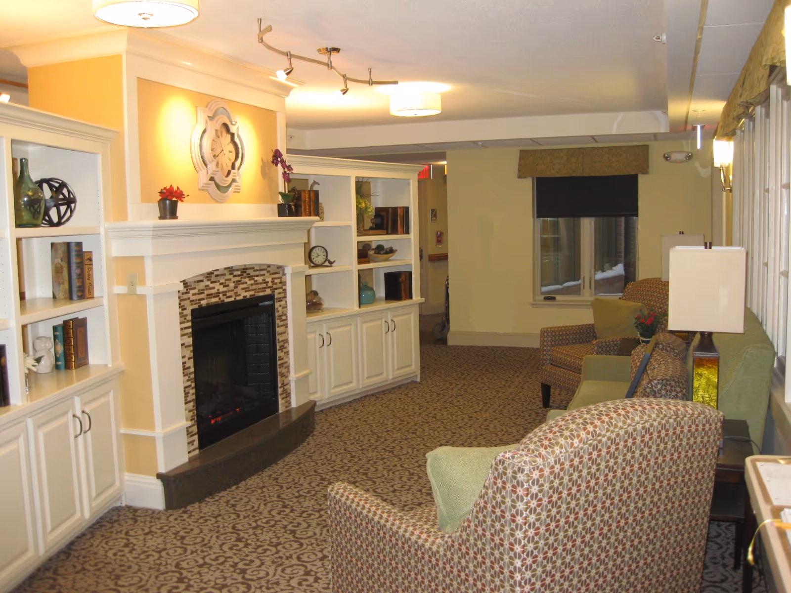 A cozy living room area with patterned carpet, upholstered armchairs, a green sofa, and a fireplace with a decorative clock above it. White built-in shelves flank the fireplace, holding books and decorative items. The room is softly lit with ceiling and wall lights, and a window with a valance is visible in the background.