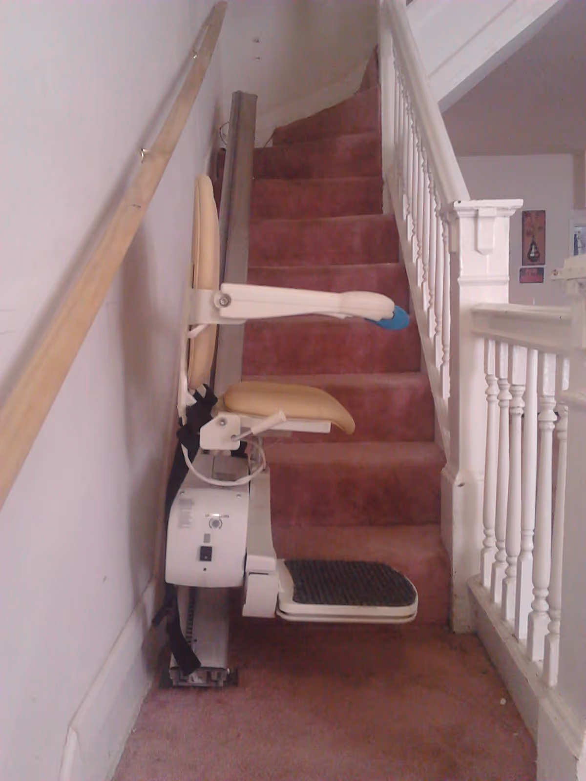 A stairlift installed on a carpeted staircase with red carpet. The stairlift has a beige seat and armrests, and a footrest with a black non-slip surface. The staircase has a wooden handrail on the left and white balusters on the right.