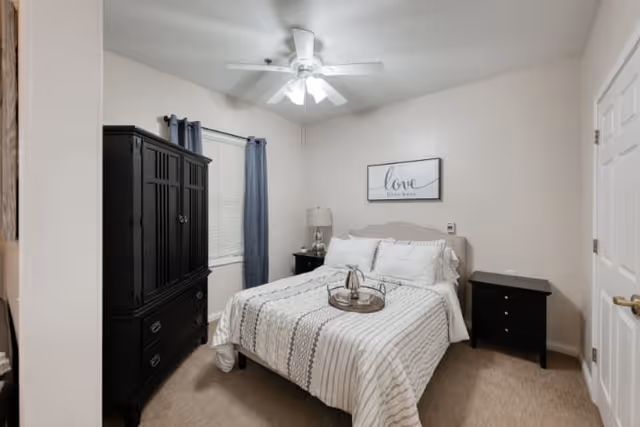 A cozy bedroom with a neatly made bed featuring white and gray striped bedding. There is a black armoire on the left side, a nightstand with a lamp next to the bed, and another small black nightstand on the right. A ceiling fan with lights is on the ceiling, and a framed sign above the bed reads 'love lives here'. The walls are light-colored, and there is a window with dark curtains behind the armoire.