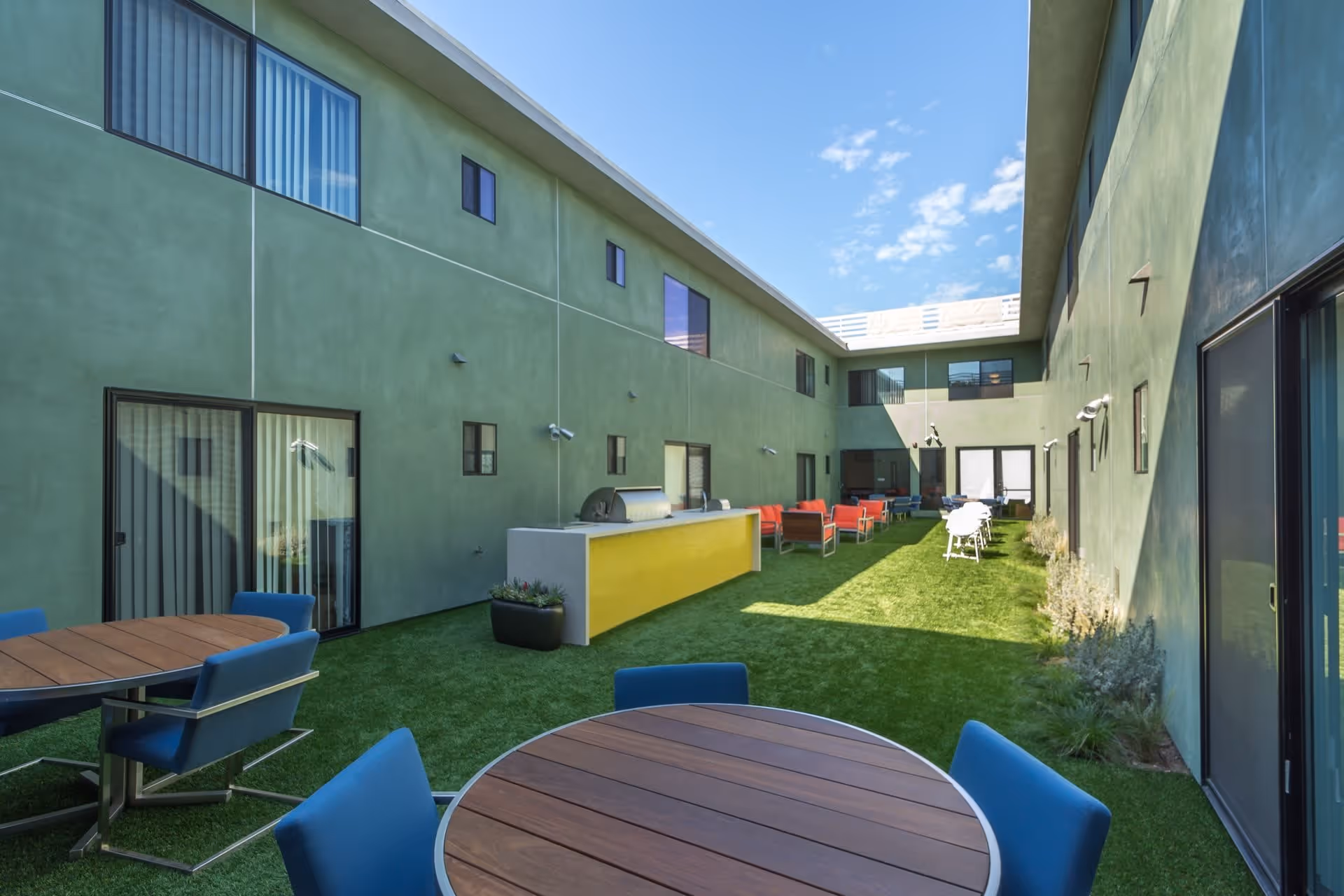 Outdoor courtyard area of a senior living facility with green artificial grass, round wooden tables with blue chairs, a yellow outdoor grill station, and red cushioned seating along the walls of a two-story green building under a blue sky with some clouds.