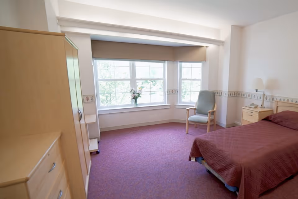 A simple, clean bedroom with a single bed covered in a maroon quilt, a wooden nightstand with a lamp and telephone, a wooden wardrobe and dresser, a green cushioned chair, and large windows with beige blinds letting in natural light. There is a vase with flowers on the windowsill and a decorative border along the walls.