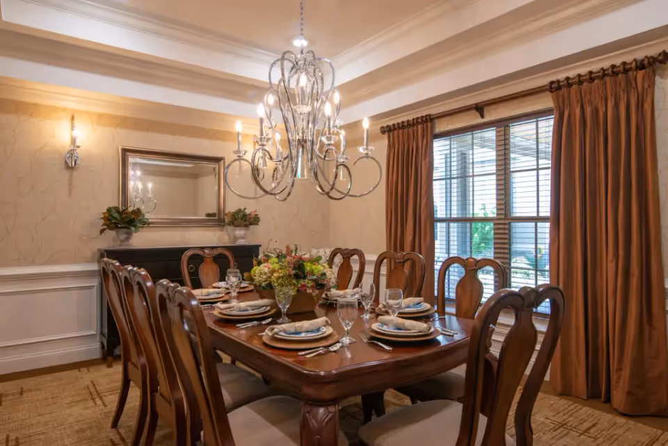 A formal dining room with a large wooden dining table set for eight people. The table is decorated with a floral centerpiece and place settings including plates, napkins, glasses, and silverware. The room features a chandelier hanging above the table, a large window with brown curtains, a mirror on the wall, and two wall sconces providing light.