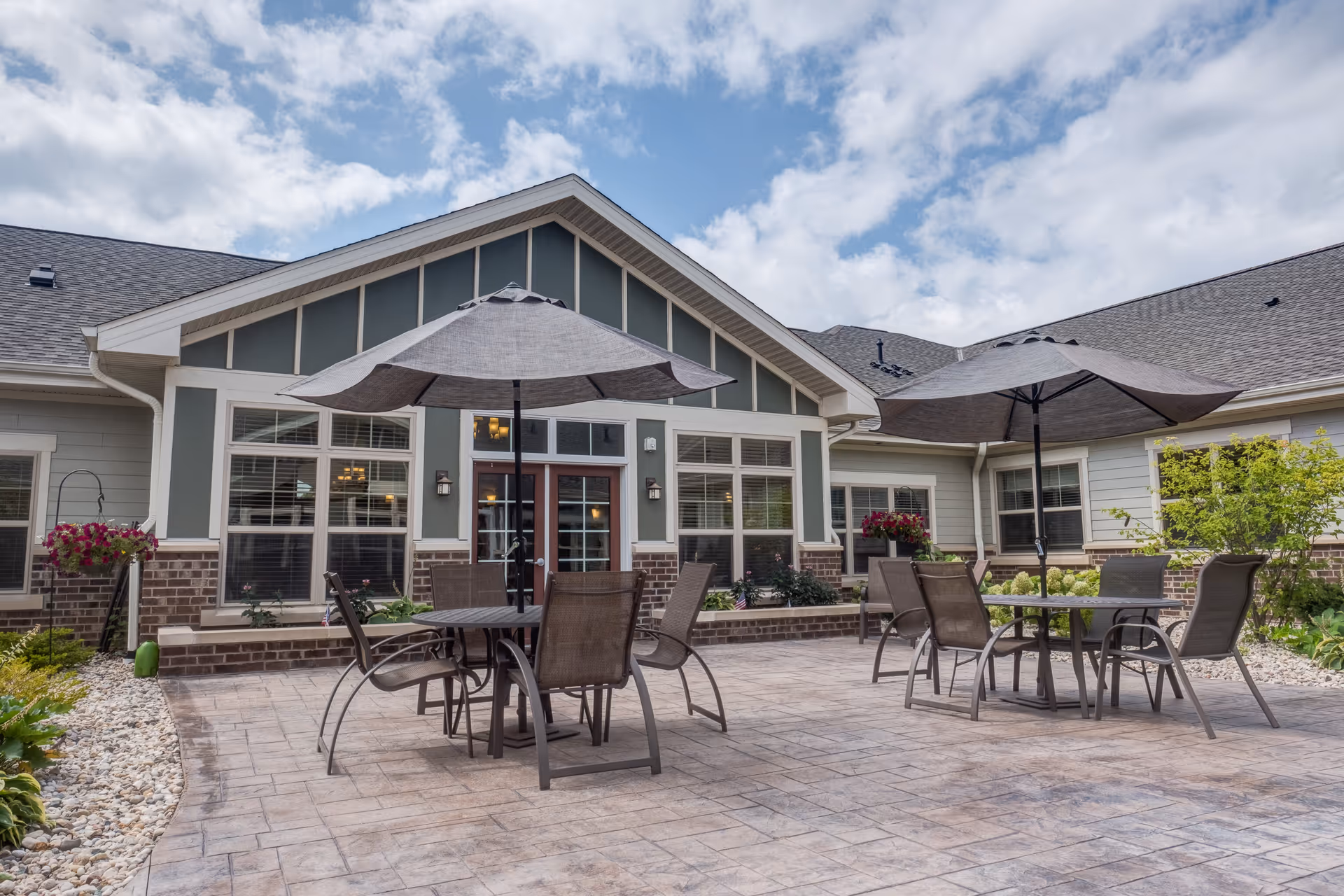 Outdoor patio area at Heritage Court Memory Care with two round tables, each shaded by a large umbrella and surrounded by chairs. The patio is paved and bordered by landscaping with flowers and greenery. The building exterior features large windows and a peaked roof under a partly cloudy sky.