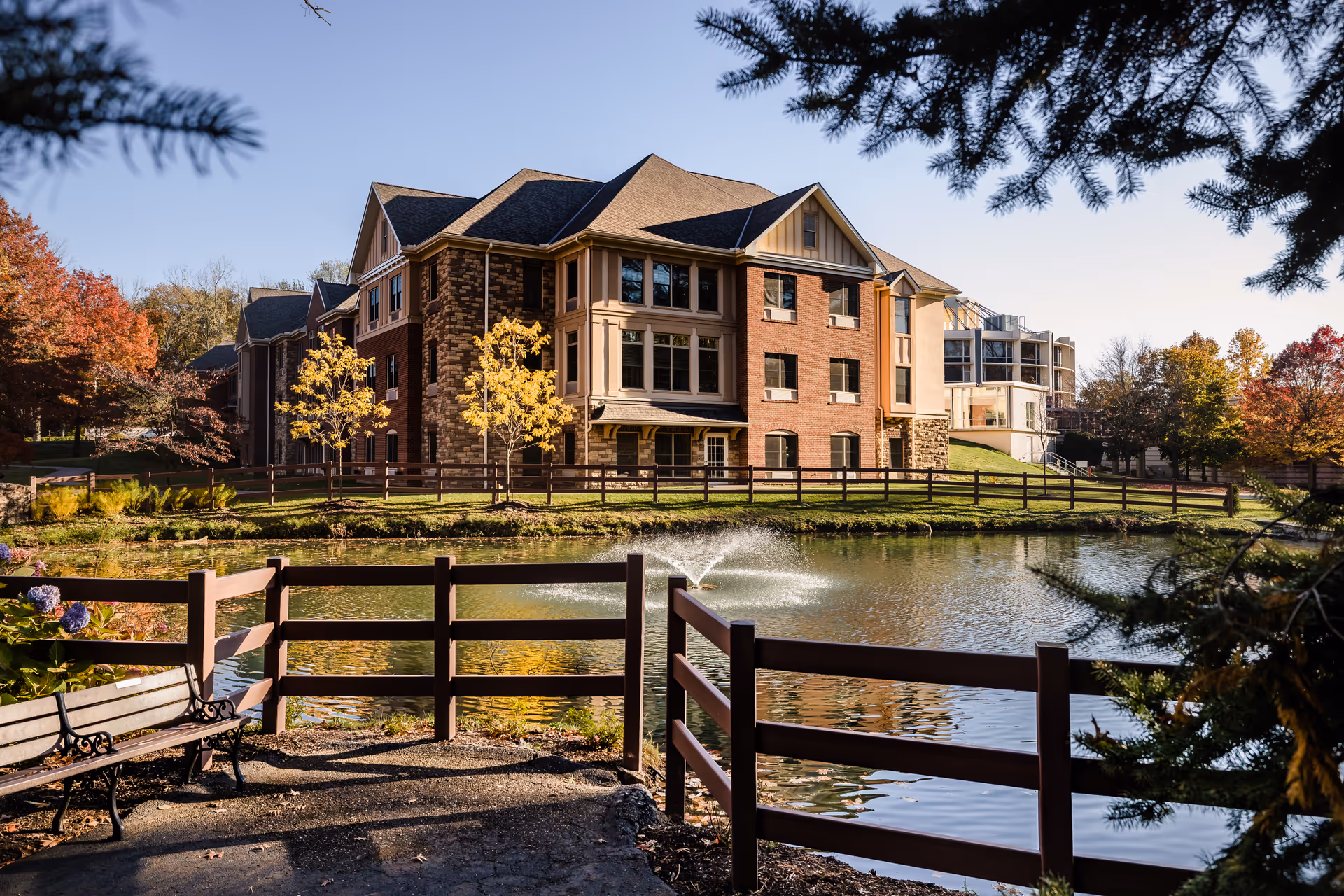 A large multi-story brick and stone building beside a pond with a water fountain, surrounded by a wooden fence and trees with autumn foliage. A bench is visible near the pond, and the scene is framed by evergreen branches.