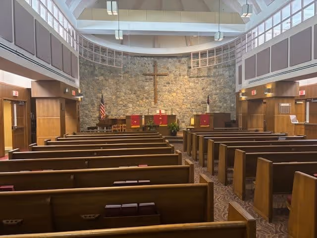 Interior view of a chapel with wooden pews arranged in rows facing a stone wall with a large wooden cross mounted on it. The chapel has a high ceiling with skylights and hanging lights. There are American and Christian flags on either side of the altar area, which is decorated with red cloth banners and plants.