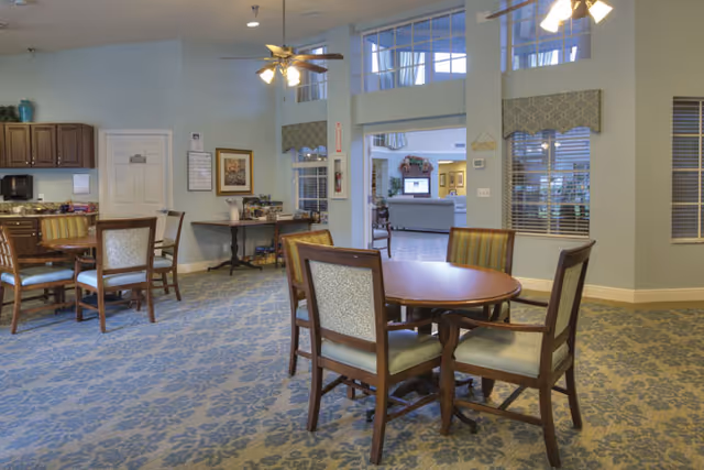 A spacious dining area in an assisted living facility with round wooden tables and cushioned chairs. The room has patterned carpet flooring, light blue walls, and large windows with valances. Ceiling fans with lights hang from the ceiling. In the background, there is a doorway leading to another room with additional seating and decor.