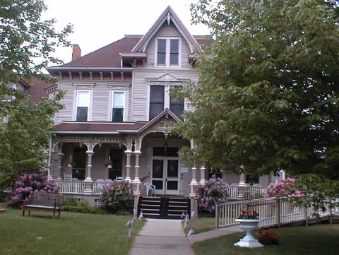 A large, two-story Victorian-style house with a steep roof and multiple windows. The house has a covered front porch with decorative columns and railings. There are trees and flowering bushes around the house, a bench on the lawn, and a paved walkway leading to the front steps. A ramp is also visible on the right side of the porch.