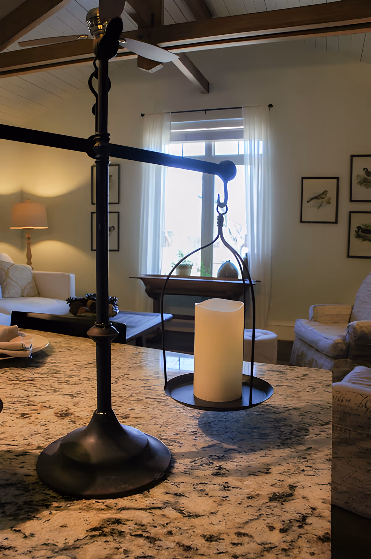 A cozy living room with a granite countertop in the foreground holding a decorative candle holder with a white candle. In the background, there is a white sofa with cushions, a wooden coffee table, a floor lamp, framed bird artwork on the wall, and a window with sheer white curtains letting in natural light.