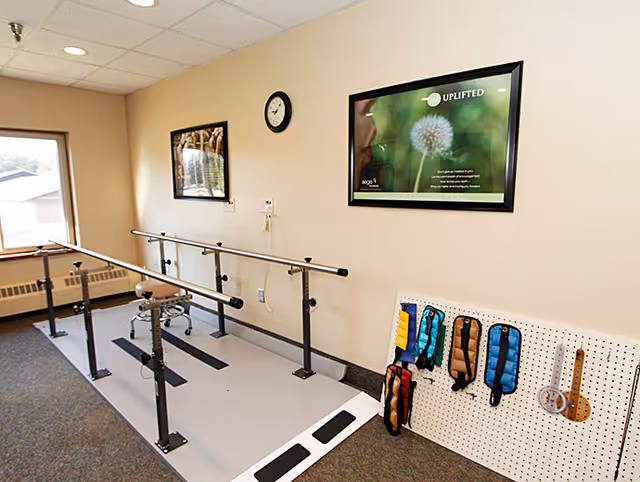 A rehabilitation or physical therapy room with parallel bars for walking exercises, a stool on wheels, a clock on the wall, framed pictures, and a pegboard holding various weighted wrist or ankle straps.
