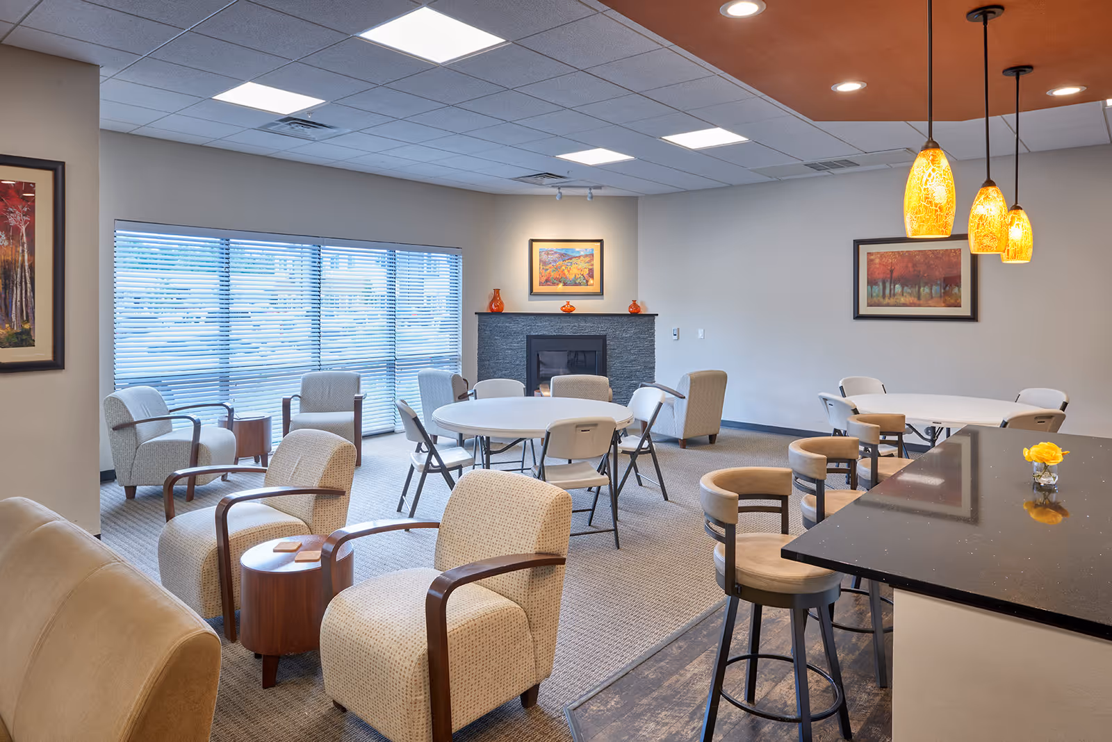 A well-lit common area with multiple seating options including cushioned armchairs, folding chairs around round tables, and bar stools at a black countertop. The room features a fireplace with decorative vases and framed artwork above it, large windows with blinds, and three hanging pendant lights with amber-colored shades.