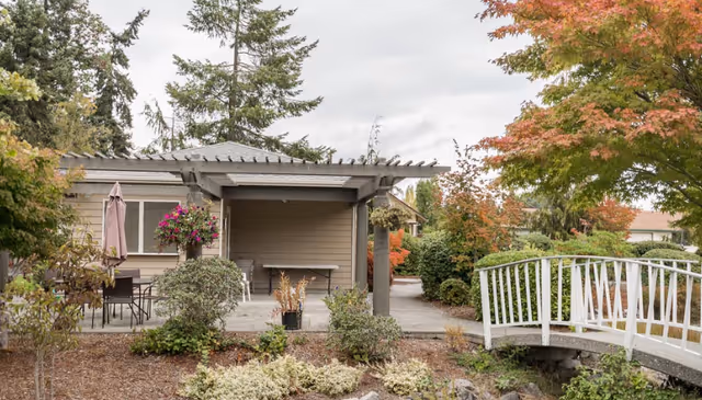 Outdoor view of a senior living facility area featuring a small covered patio with a table and chairs, hanging flower baskets, surrounding bushes and trees with autumn foliage, and a white wooden footbridge over a small landscaped area.