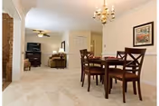 A spacious interior room featuring a dining area with a wooden table and four chairs under a chandelier. In the background, there is a living area with a sofa, armchair, television, and ceiling fan. The room has light-colored carpet flooring and neutral-colored walls with framed artwork.