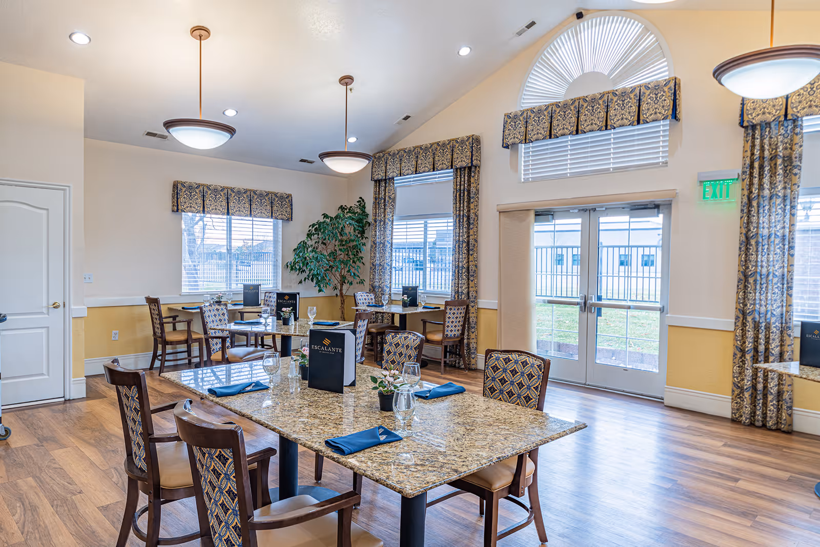 Bright communal dining room with several set tables and chairs, large windows with patterned curtains, and double glass doors.