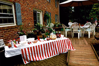 Outdoor patio area with a brick wall and windows in the background. A table covered with a red and white striped tablecloth is set with various food items and drinks. Several white plastic chairs and tables are arranged on a wooden deck nearby, surrounded by greenery.