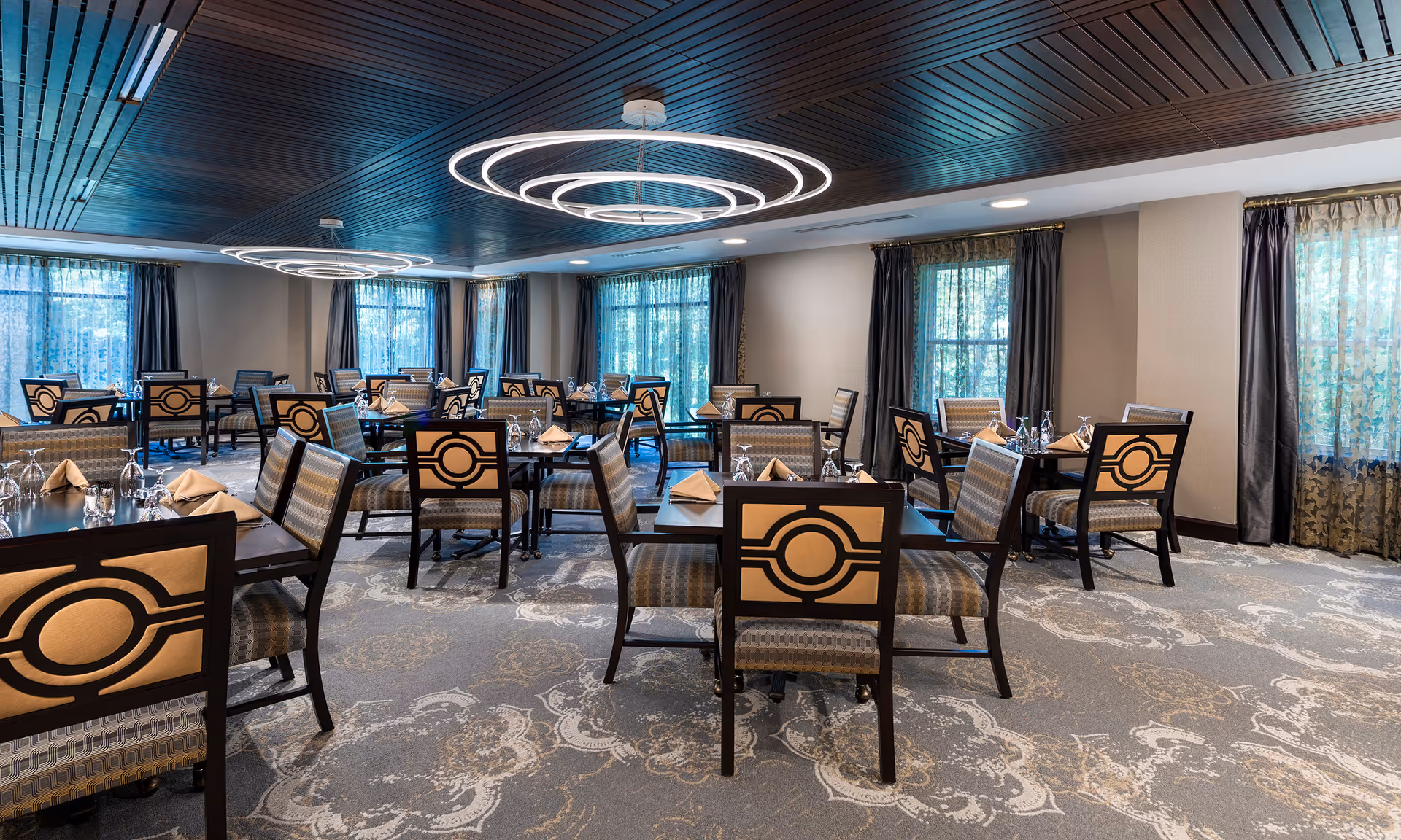 A spacious dining room with multiple tables and chairs arranged neatly. Each table is set with folded napkins and upside-down glasses. The room features large windows with sheer and dark curtains, a patterned carpet, and modern circular ceiling lights with a wooden ceiling panel design.