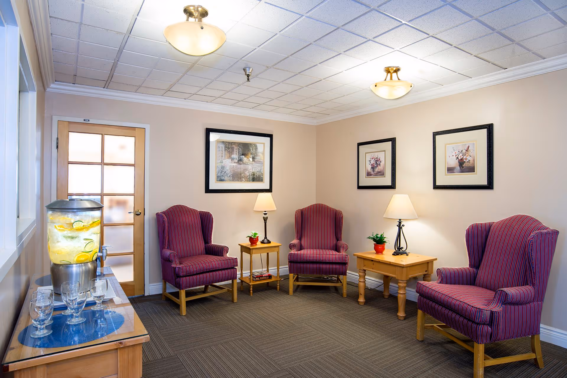 A bright sitting room with four striped wingback chairs arranged around wooden side tables and lamps, framed artwork on the walls, and a lemon water dispenser on a console.