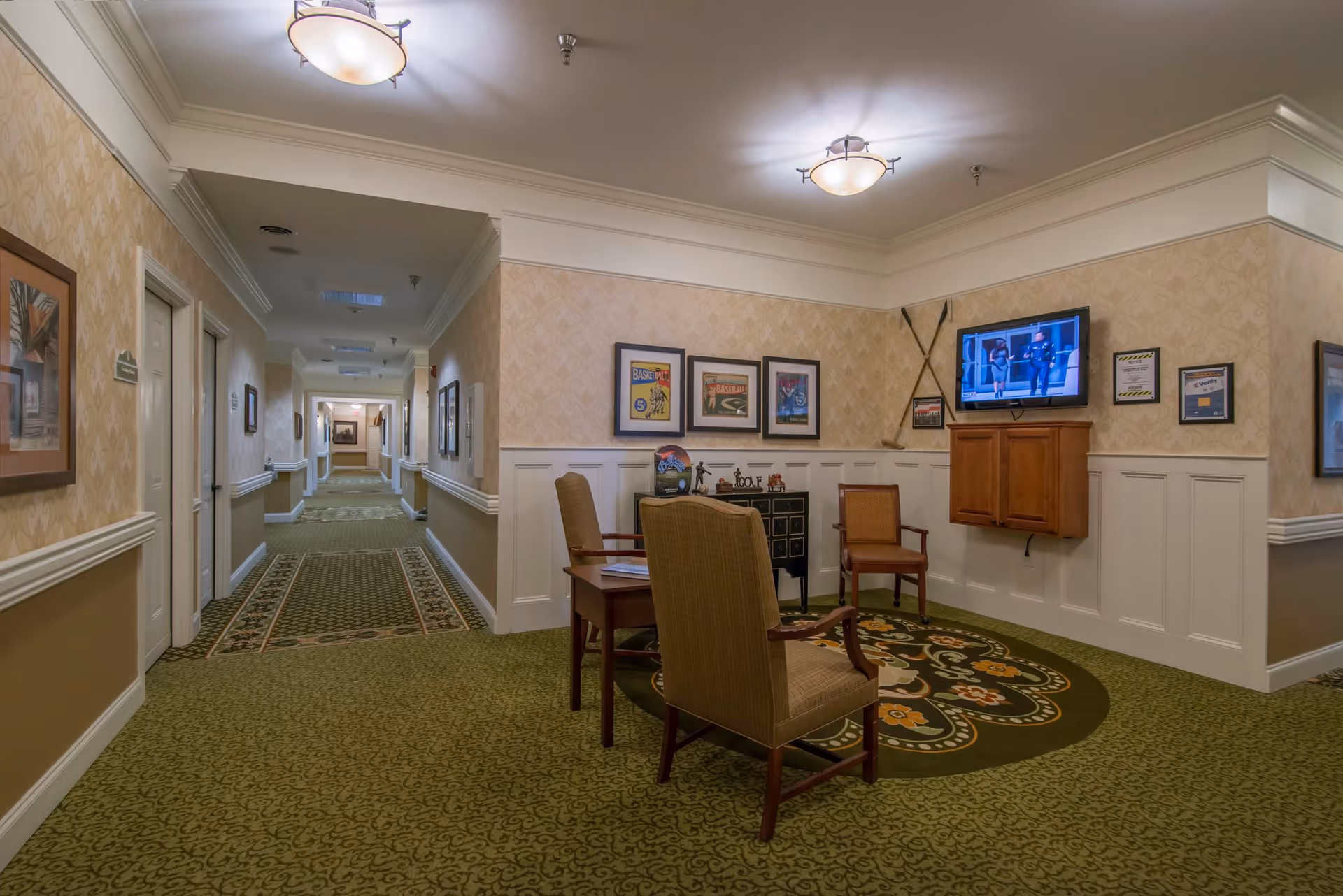 A hallway in a senior living facility with patterned green carpet and beige wallpaper. There is a small seating area with two chairs and a table on a circular rug. The wall has framed pictures, a mounted TV, and decorative items including crossed baseball bats. The hallway extends into the distance with doors and framed pictures along the walls.