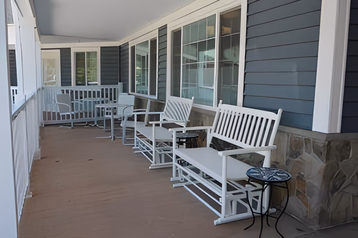A covered outdoor porch area with white wooden rocking benches and chairs, small tables, and large windows along the building wall. The porch has a brown floor and blue siding with white trim on the building exterior.