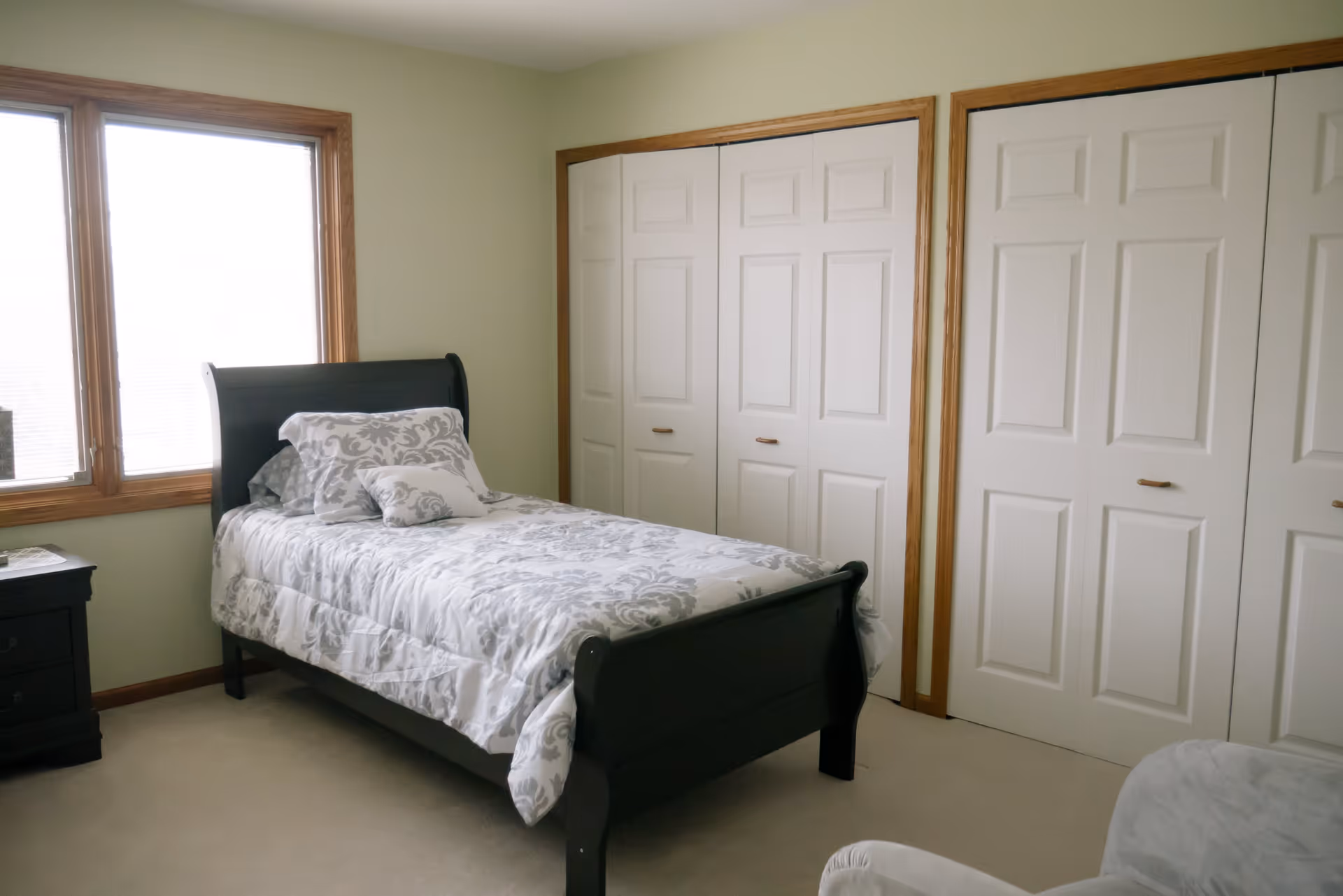 A bright bedroom with a single dark wooden bed featuring white and gray patterned bedding and pillows. There is a window with wooden trim on the left side, a dark wooden nightstand with drawers next to the bed, and two white closets with wooden trim on the right wall. The room has light-colored walls and carpeted flooring.
