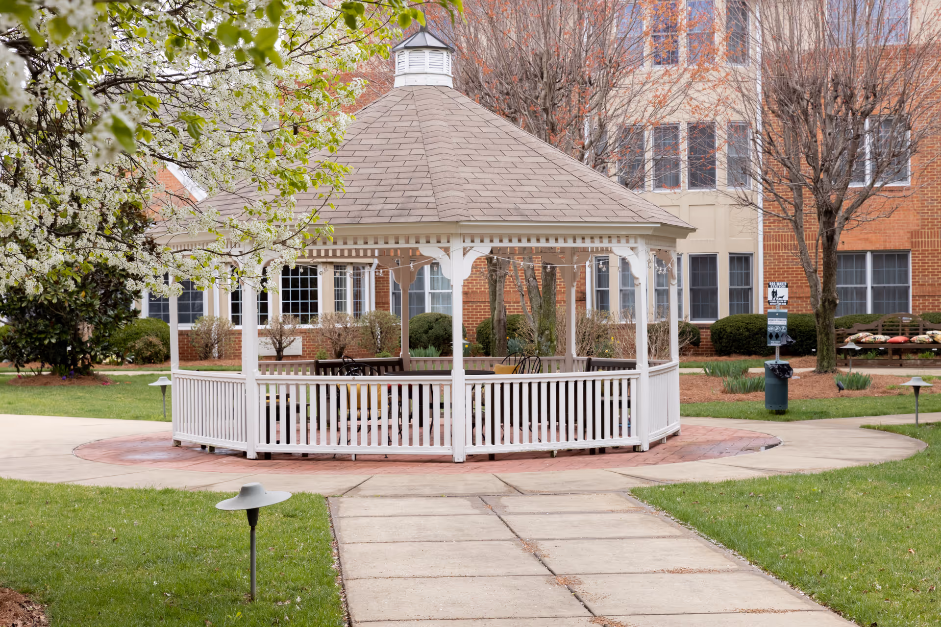 A white wooden gazebo sits in a landscaped courtyard in front of a brick senior living building.